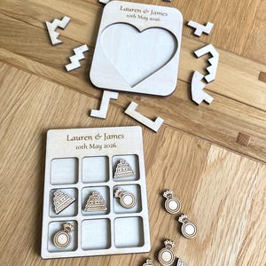 K&ouml;nnte beinhalten: Ein helles Holz-Tic-Tac-Toe-Spielbrett und ein herzf&ouml;rmiger Puzzlerahmen, beide graviert mit dem Text Lauren & James 10th May 2026. Das Set enth&auml;lt kleine Holzspielsteine in Form von Hochzeitstorten und Verlobungsringen.