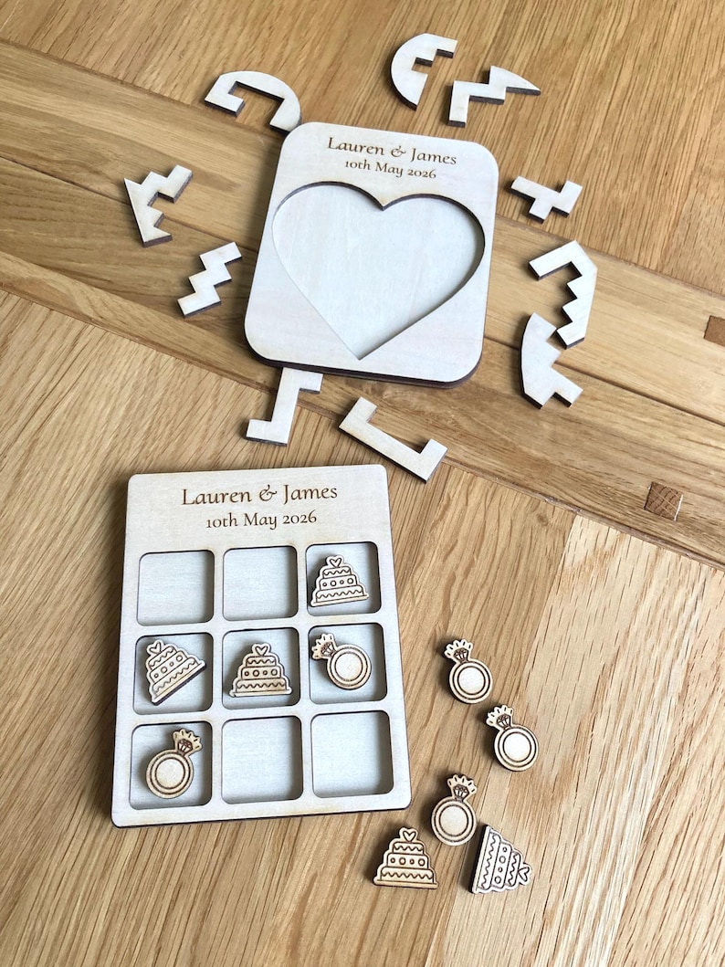 K&ouml;nnte beinhalten: Ein helles Holz-Tic-Tac-Toe-Spielbrett und ein herzf&ouml;rmiger Puzzlerahmen, beide graviert mit dem Text Lauren & James 10th May 2026. Das Set enth&auml;lt kleine Holzspielsteine in Form von Hochzeitstorten und Verlobungsringen.
