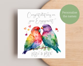 Könnte beinhalten: Eine weiße Karte mit einer Aquarell-Illustration von zwei bunten Vögeln, die auf einem Ast sitzen. Die Vögel sind von kleinen roten Herzen umgeben. Der Text "Congratulations on your Engagement" ist in schwarzer Kursivschrift über den Vögeln geschrieben. Die Namen "Katie & Beth" sind in schwarzer Kursivschrift unter den Vögeln geschrieben.