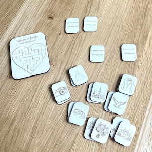 K&ouml;nnte beinhalten: Ein Set aus hellen Holz-Puzzleteilen und rechteckigen Pl&auml;ttchen mit Hochzeitsmotiven. Das gr&ouml;&szlig;te Teil zeigt ein herzf&ouml;rmiges Labyrinth und den Text Lauren & James 10th May 2026. Kleinere Pl&auml;ttchen zeigen eingravierte Symbole wie Kamera, Kleid, Kirche, Ringe, Taube, Blumenstrau&szlig; und Geschenk.