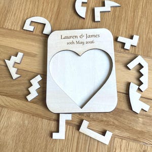 K&ouml;nnte beinhalten: Ein heller Holz-Puzzle-Rahmen mit einem herzf&ouml;rmigen Ausschnitt in der Mitte. Der obere Teil des Rahmens ist mit dem Text Lauren & James 10th May 2026 graviert. Mehrere unregelm&auml;&szlig;ige Holz-Puzzleteile sind um den Rahmen angeordnet.