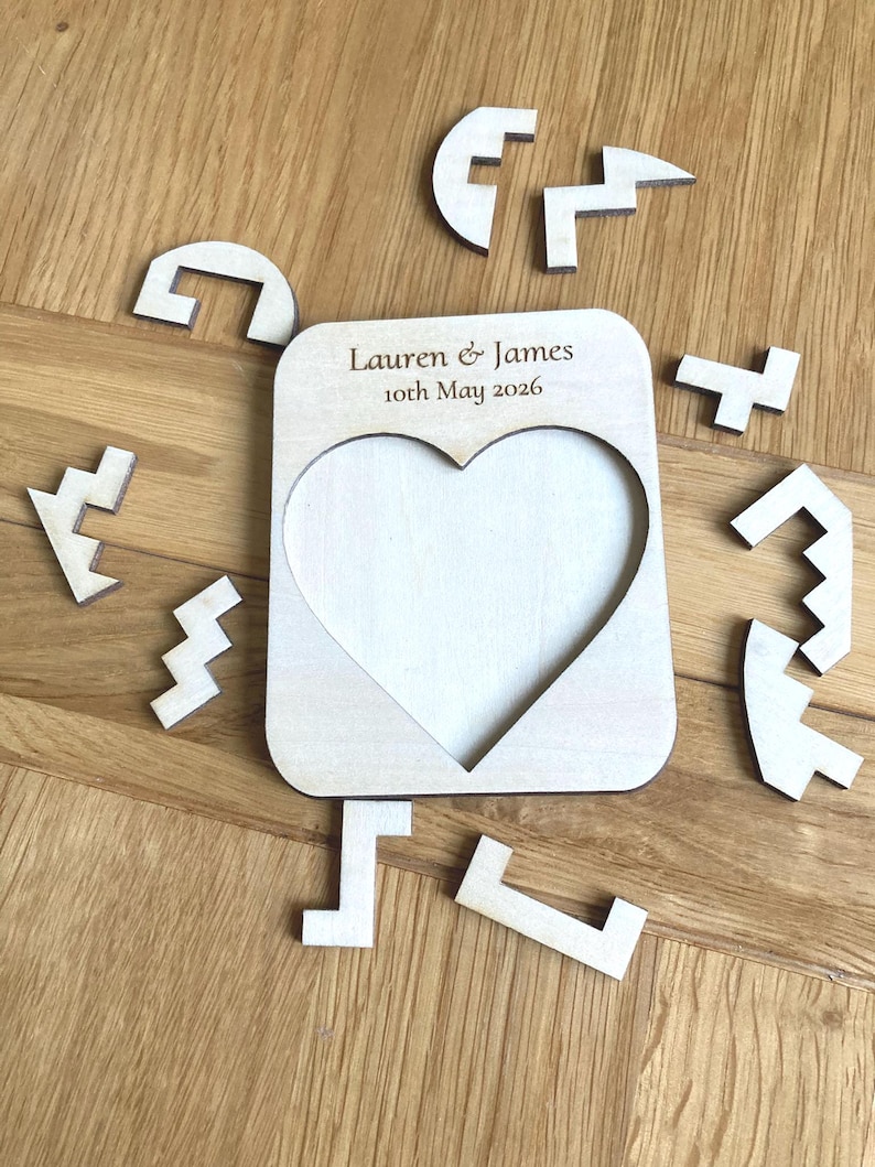 K&ouml;nnte beinhalten: Ein heller Holz-Puzzle-Rahmen mit einem herzf&ouml;rmigen Ausschnitt in der Mitte. Der obere Teil des Rahmens ist mit dem Text Lauren & James 10th May 2026 graviert. Mehrere unregelm&auml;&szlig;ige Holz-Puzzleteile sind um den Rahmen angeordnet.