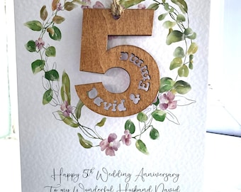 Könnte beinhalten: Eine weiße Grußkarte mit einem Aquarellkranz aus grünen Blättern und kleinen violetten Blüten. Eine hölzerne Zahl 5 ist mit einer Schnur befestigt und mit den Namen David & Emma graviert. Der Text lautet: Happy 5th Wedding Anniversary To my Wonderful Husband David.