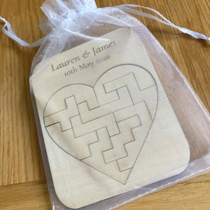 K&ouml;nnte beinhalten: Ein helles, herzf&ouml;rmiges Holzpuzzle in einem durchsichtigen wei&szlig;en Organzabeutel mit Kordelzug. Das Holz zeigt ein lasergraviertes Labyrinth-Muster und den Text Lauren & James 10th May 2026 in einer dunklen Serifenschrift.