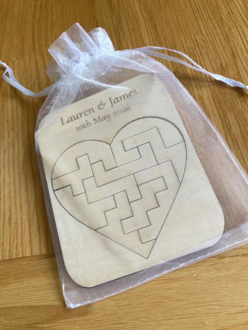 K&ouml;nnte beinhalten: Ein helles, herzf&ouml;rmiges Holzpuzzle in einem durchsichtigen wei&szlig;en Organzabeutel mit Kordelzug. Das Holz zeigt ein lasergraviertes Labyrinth-Muster und den Text Lauren & James 10th May 2026 in einer dunklen Serifenschrift.