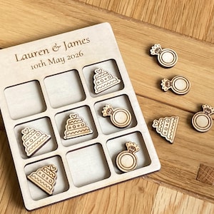 K&ouml;nnte beinhalten: Ein helles Holzkreuz-und-Null-Spielbrett mit der Gravur Lauren & James 10th May 2026. Das Brett verf&uuml;gt &uuml;ber ein Raster mit Holzspielfiguren in Form von mehrst&ouml;ckigen Hochzeitstorten und Diamantverlobungsringen.