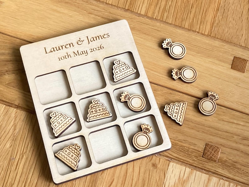 K&ouml;nnte beinhalten: Ein helles Holzkreuz-und-Null-Spielbrett mit der Gravur Lauren & James 10th May 2026. Das Brett verf&uuml;gt &uuml;ber ein Raster mit Holzspielfiguren in Form von mehrst&ouml;ckigen Hochzeitstorten und Diamantverlobungsringen.