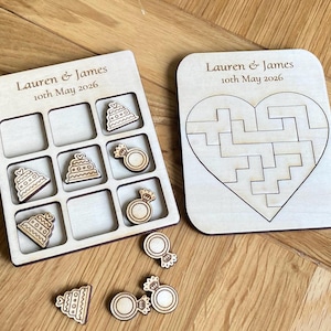 K&ouml;nnte beinhalten: Ein helles Holzkreuz-und-Null-Spielbrett und ein herzf&ouml;rmiges Puzzlebrett. Beide tragen den Text Lauren & James 10th May 2026. Das Spielbrett enth&auml;lt h&ouml;lzerne Spielfiguren in Form von Hochzeitstorten und Ringen.