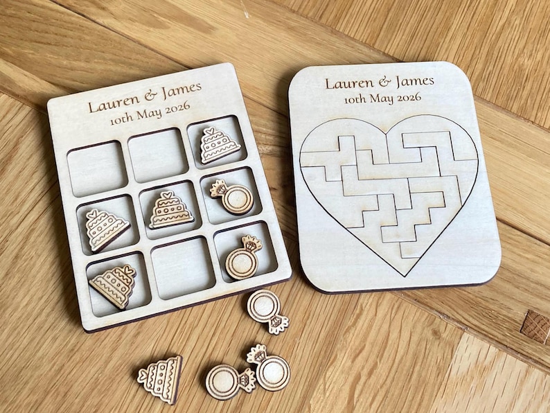 K&ouml;nnte beinhalten: Ein helles Holzkreuz-und-Null-Spielbrett und ein herzf&ouml;rmiges Puzzlebrett. Beide tragen den Text Lauren & James 10th May 2026. Das Spielbrett enth&auml;lt h&ouml;lzerne Spielfiguren in Form von Hochzeitstorten und Ringen.