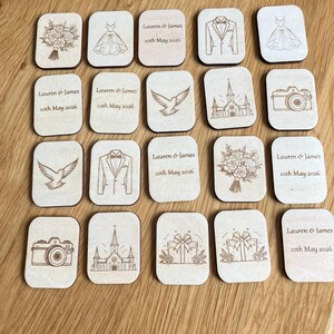 K&ouml;nnte beinhalten: Eine Sammlung rechteckiger Holzpl&auml;ttchen mit lasergravierten Hochzeitssymbolen wie Blumenstrau&szlig;, Kleid, Smoking, Taube, Kirche, Kamera und Geschenk. Einige Pl&auml;ttchen sind mit dem Text Lauren & James 10th May 2026 personalisiert.