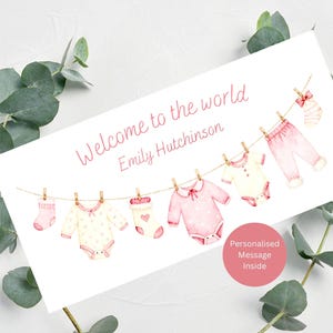 May include: A greeting card with the text "Welcome to the world" and a baby's name. The card features watercolor illustrations of baby clothes hanging on a clothesline, in shades of pink and cream.