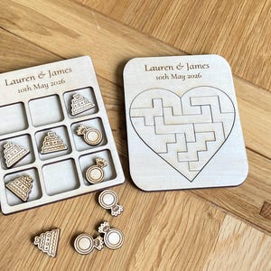 K&ouml;nnte beinhalten: Ein helles Holzspielbrett f&uuml;r Tic-Tac-Toe mit Spielfiguren in Form von Torten und Ringen sowie ein herzf&ouml;rmiges Puzzlebrett. Beide Teile sind mit dem Text Lauren & James 10th May 2026 graviert. Die Spielfiguren weisen detaillierte Gravuren auf.
