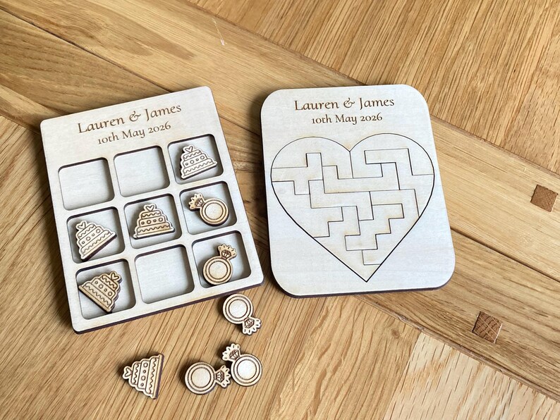 K&ouml;nnte beinhalten: Ein helles Holzspielbrett f&uuml;r Tic-Tac-Toe mit Spielfiguren in Form von Torten und Ringen sowie ein herzf&ouml;rmiges Puzzlebrett. Beide Teile sind mit dem Text Lauren & James 10th May 2026 graviert. Die Spielfiguren weisen detaillierte Gravuren auf.