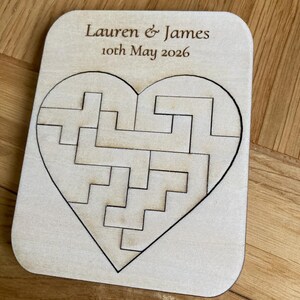 K&ouml;nnte beinhalten: Eine rechteckige, helle Holzplatte mit einem herzf&ouml;rmigen Puzzle aus ineinandergreifenden Teilen. Der Text "Lauren & James" und "10th May 2026" ist in dunkelbrauner Schrift oben auf der Holzoberfl&auml;che eingraviert.