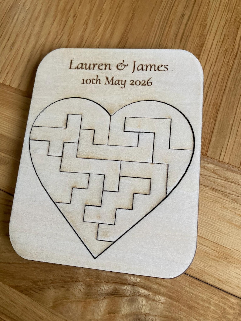 K&ouml;nnte beinhalten: Eine rechteckige, helle Holzplatte mit einem herzf&ouml;rmigen Puzzle aus ineinandergreifenden Teilen. Der Text "Lauren & James" und "10th May 2026" ist in dunkelbrauner Schrift oben auf der Holzoberfl&auml;che eingraviert.