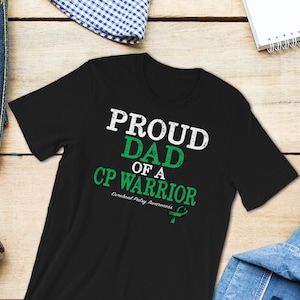 Op de afbeelding: Zwart T-shirt met witte tekst die "Proud Dad of a CP Warrior" luidt met een groen lint eronder. Bewustwording van Cerebral Palsy.