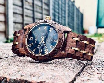 Puede incluir: Un reloj de pulsera de madera marrón con una esfera negra y detalles dorados. El reloj tiene marcadores de hora en números romanos y una ventana de fecha en la posición de las 3 en punto. La correa del reloj está hecha de madera y tiene un cierre de metal dorado.
