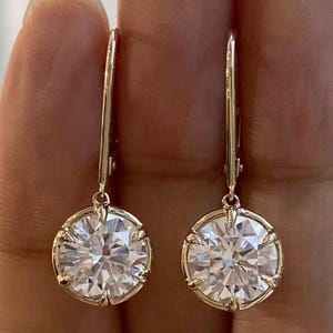May include: A pair of gold drop earrings featuring large, round, clear gemstones. The gemstones are set in a gold bezel setting, and the earrings have a simple, elegant design. The earrings are shown against a neutral background.