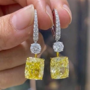 May include: A pair of drop earrings featuring large, cushion-cut yellow stones suspended from smaller, round, clear stones. The earring hooks are encrusted with small, clear, pavé-set stones, creating a bright, reflective, and multi-layered appearance.