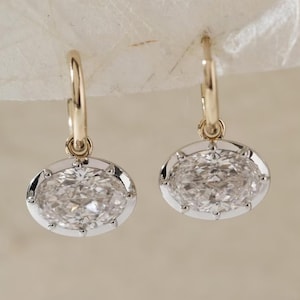 May include: A pair of gold hoop earrings with oval-shaped, clear gemstones. The gemstones are set in a silver-colored bezel with small, decorative prongs. The earrings are displayed against a neutral background.