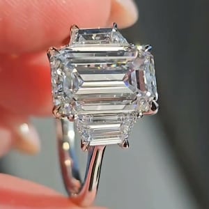 Puede incluir: Anillo de plata con un gran diamante rectangular con talla esmeralda como pieza central, flanqueado por dos diamantes trapezoidales más pequeños. La banda del anillo es de plata pulida y los diamantes brillan.