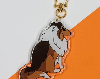 Op de afbeelding: Een heldere acryl sleutelhanger met een cartoon collie hond met bruin, wit en bruin haar. De hond zit en de sleutelhanger heeft een gouden stervormige sluiting. De achtergrond is wit en oranje.