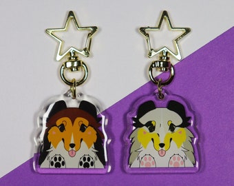 Op de afbeelding: Twee acryl sleutelhangers met cartoonhonden. De sleutelhanger aan de linkerkant heeft een bruine en witte hond met een roze tong die uitsteekt. De sleutelhanger aan de rechterkant heeft een zwarte en witte hond met een geel en wit vachtpatroon en een roze tong die uitsteekt. Beide sleutelhangers hebben een gouden stervormige hanger die aan een gouden ring is bevestigd.
