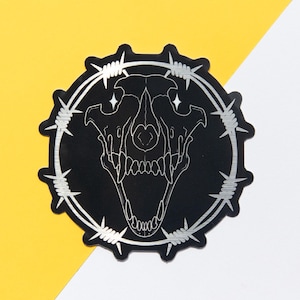 May include: A black sticker featuring a detailed illustration of a skull within a circle of barbed wire. The skull has visible teeth and eye sockets. The sticker is set against a yellow and white background.