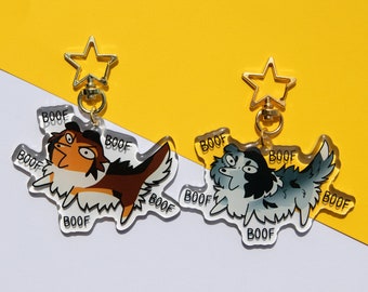 Op de afbeelding: Twee acryl sleutelhangers met cartoon-honden met het woord "BOOF" gedrukt rond de randen. De honden zijn beide wit met bruine en zwarte markeringen. Eén hond is bruin en de andere is grijs.