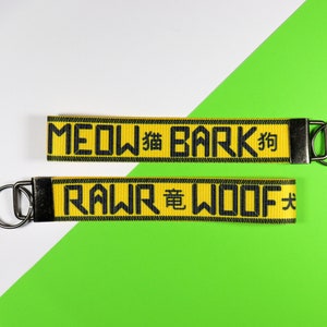 Pode incluir: Dois chaveiros amarelos com texto preto e ferragens met&aacute;licas. O chaveiro superior diz "MEOW" e "BARK" com caracteres chineses. O chaveiro inferior diz "RAWR" e "WOOF" com caracteres chineses. O fundo &eacute; branco e verde.