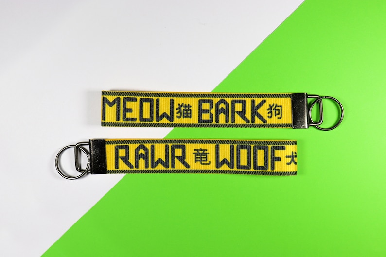 Pode incluir: Dois chaveiros amarelos com texto preto e ferragens met&aacute;licas. O chaveiro superior diz "MEOW" e "BARK" com caracteres chineses. O chaveiro inferior diz "RAWR" e "WOOF" com caracteres chineses. O fundo &eacute; branco e verde.