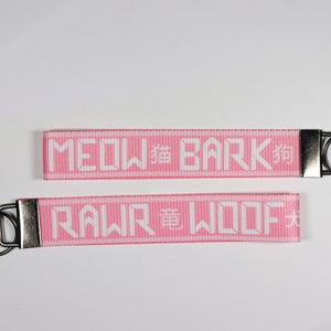 Pode incluir: Dois chaveiros rosa com texto branco. O chaveiro superior diz "MEOW" e "BARK" com um caractere japon&ecirc;s. O chaveiro inferior diz "RAWR" e "WOOF" com um caractere japon&ecirc;s. Cada chaveiro tem um fecho e um anel de metal prateado.