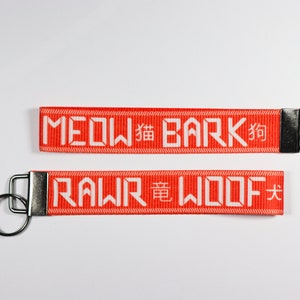 Pode incluir: Dois chaveiros vermelhos com texto branco. O chaveiro superior diz "MEOW 貓 BARK 狗" e o chaveiro inferior diz "RAWR 竜 WOOF 犬". Cada chaveiro tem um anel de metal prateado.