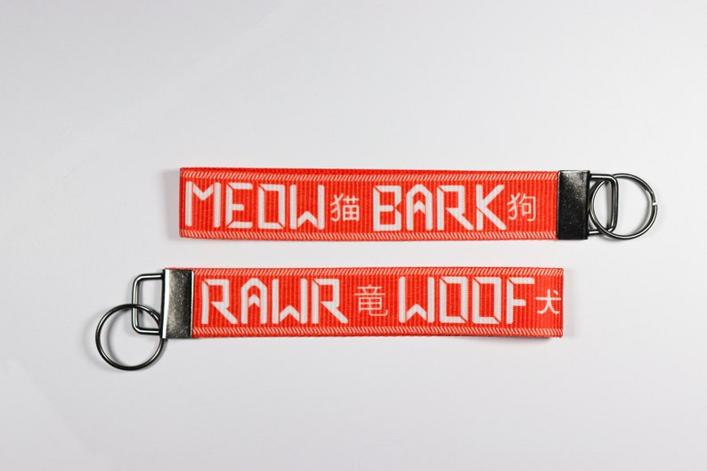 Pode incluir: Dois chaveiros vermelhos com texto branco. O chaveiro superior diz "MEOW 貓 BARK 狗" e o chaveiro inferior diz "RAWR 竜 WOOF 犬". Cada chaveiro tem um anel de metal prateado.