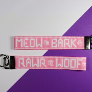 Pode incluir: Dois chaveiros rosa com texto branco. Um diz "MEOW 貓 BARK 狗" e o outro "RAWR 龍 WOOF 犬". Cada chaveiro tem um fecho de metal prateado e um anel de metal preto. O fundo &eacute; branco e roxo.