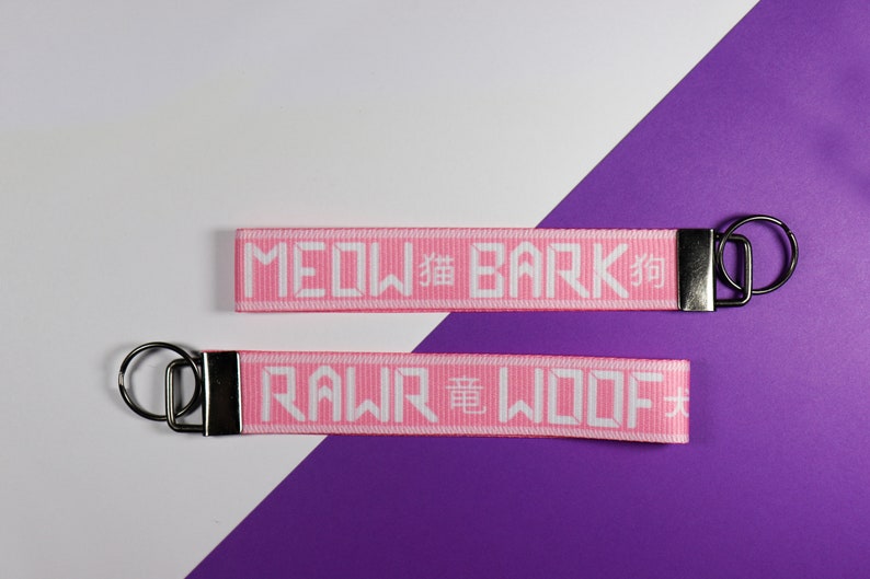 Pode incluir: Dois chaveiros rosa com texto branco. Um diz "MEOW 貓 BARK 狗" e o outro "RAWR 龍 WOOF 犬". Cada chaveiro tem um fecho de metal prateado e um anel de metal preto. O fundo &eacute; branco e roxo.