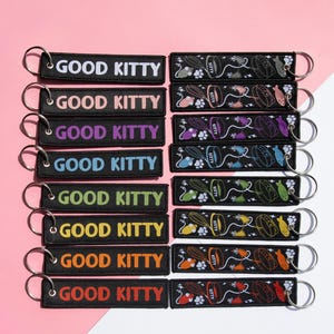 May include: A selection of black keyrings, each with the words "GOOD KITTY" in different colours. The keyrings have a metal ring and a design of cat-related items such as food bowls and fish.