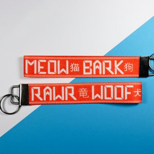 Pode incluir: Dois chaveiros laranja com texto branco e argolas prateadas. O chaveiro superior diz "MEOW 貓 BARK 狗", e o chaveiro inferior diz "RAWR 竜 WOOF 犬". O fundo &eacute; dividido diagonalmente, com se&ccedil;&otilde;es brancas e azuis.