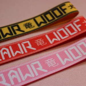 Pode incluir: Tr&ecirc;s chaveiros coloridos, amarelo, vermelho e rosa, com as palavras "RAWR WOOF" em letras brancas e caracteres chineses. Os chaveiros s&atilde;o feitos de tecido tecido.