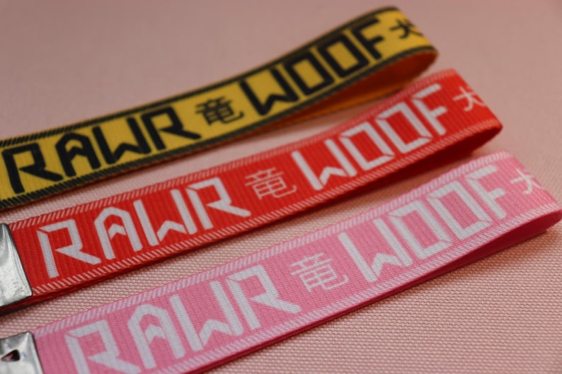 Pode incluir: Tr&ecirc;s chaveiros coloridos, amarelo, vermelho e rosa, com as palavras "RAWR WOOF" em letras brancas e caracteres chineses. Os chaveiros s&atilde;o feitos de tecido tecido.