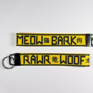 Pode incluir: Dois chaveiros amarelos com texto preto e argolas prateadas. O chaveiro superior diz "MEOW 猫 BARK 狗" e o chaveiro inferior diz "RAWR 竜 WOOF 犬". Os chaveiros t&ecirc;m uma borda preta.
