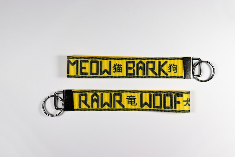 Pode incluir: Dois chaveiros amarelos com texto preto e argolas prateadas. O chaveiro superior diz "MEOW 猫 BARK 狗" e o chaveiro inferior diz "RAWR 竜 WOOF 犬". Os chaveiros t&ecirc;m uma borda preta.