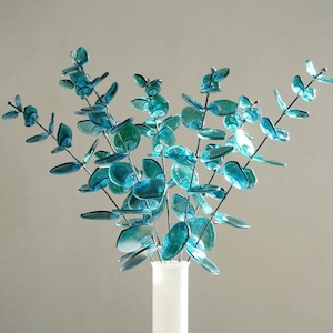 May include: A white, round vase holds a collection of faux eucalyptus branches. The leaves are a vibrant teal color, with a glossy finish. The vase has a smooth, matte finish and a long neck. The arrangement is set against a neutral background.
