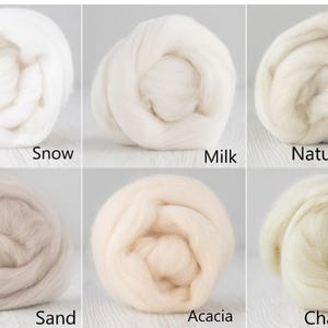 Op de afbeelding: Zes ronde bundels van zachte, vezelige wolvilt, gerangschikt in een raster. De kleuren zijn gelabeld als Snow, Milk, Natural White, Sand, Acacia en Champagne. Elke bundel vertoont een pluizige textuur en een unieke tint wit, cr&egrave;me of beige.