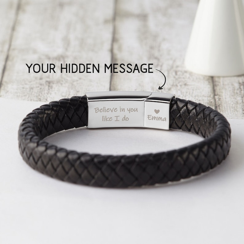 May include: Black leather bracelet with a silver clasp. The clasp has an engraved message that reads "Believe in you like I do Emma" with a heart symbol.