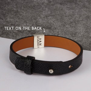May include: Black leather bracelet with a silver clasp. The bracelet has a textured exterior and a tan interior. The clasp is engraved with the initials "A & M" and the date "2/14/2017". The text "TEXT ON THE BACK" is above the clasp.