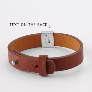 May include: A brown leather bracelet with a silver clasp. The clasp is engraved with the word "MOM" and the years "1945-2017". The bracelet has a buckle closure and a silver stud detail. The text "TEXT ON THE BACK" is above the bracelet.