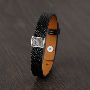 May include: Black leather bracelet with a silver square fingerprint design. The bracelet has a textured exterior and a tan interior. The clasp is a buckle style.