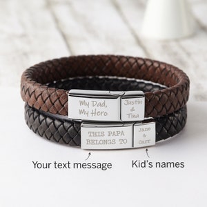 May include: Two leather bracelets with silver metal clasps. The top bracelet is brown and the bottom bracelet is black. The silver clasps are engraved with the text "My Dad, My Hero", "Justin & Tina", "THIS PAPA BELONGS TO", and "Jane & Carr".