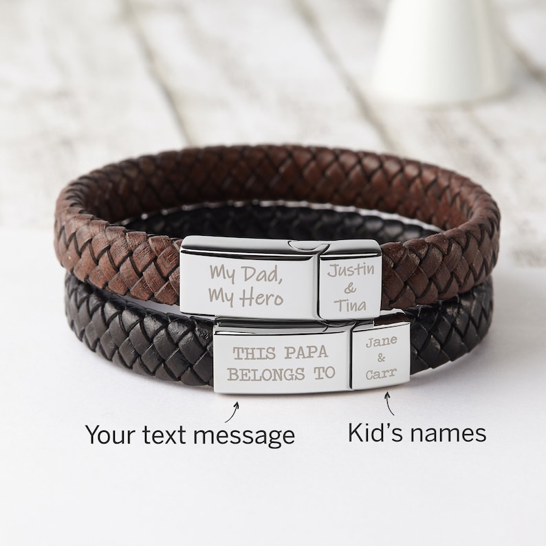 May include: Two leather bracelets with silver metal clasps. The top bracelet is brown and the bottom bracelet is black. The silver clasps are engraved with the text "My Dad, My Hero", "Justin & Tina", "THIS PAPA BELONGS TO", and "Jane & Carr".